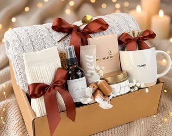 May include: A gift basket filled with a white cable-knit blanket, socks, a mug with "mom" printed on it, a candle, a bottle of room spray, and other self-care items. The basket is tied with a burgundy ribbon.