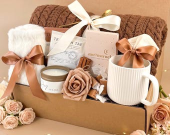 May include: A gift box filled with a white mug, a brown knit blanket, a white fluffy towel, tea packets, a candle, and a decorative rose. The box is tied with a white ribbon and a brown ribbon. The box is decorated with flowers and fairy lights.