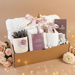 Pu&ograve; includere: Un cesto regalo pieno di una coperta bianca lavorata a maglia, una pianta succulenta in un vaso decorativo, bustine di t&egrave;, caramelle al sale, una tazza e una tavoletta di cioccolato. Il cesto &egrave; legato con un nastro rosa e si trova in una scatola di cartone marrone.