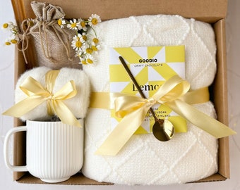 May include: A white blanket with a yellow ribbon and a small box of lemon flavored craft chocolate. The box has a gold spoon and the words "Goodio Craft Chocolate" and "Lemon" on it. The blanket is in a brown box with a white mug, a small bag of dried flowers, and a white fluffy ball with a yellow ribbon.