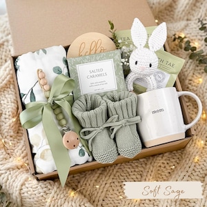 May include: A gift box filled with baby items in soft sage green and cream. Includes a blanket, booties, pacifier, bunny toy, mug with "mom" text, and salted caramels. The box is presented on a cozy, cream-colored blanket.