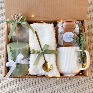 May include: A gift box filled with a green candle, a white mug, a white fluffy blanket, a small burlap bag, and a bag of sea salt caramels. The box is decorated with green and white ribbon and a sprig of greenery.