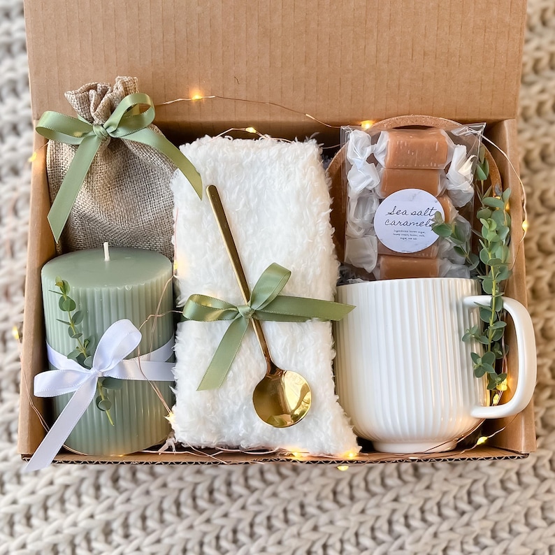May include: A gift box filled with a green candle, a white mug, a white fluffy blanket, a small burlap bag, and a bag of sea salt caramels. The box is decorated with green and white ribbon and a sprig of greenery.