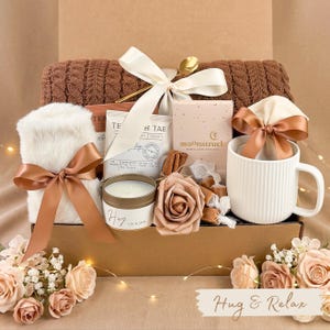 Puede incluir: Una caja de regalo con una taza blanca, una bufanda de piel sint&eacute;tica, bolsitas de t&eacute;, una vela y una rosa decorativa. La caja est&aacute; atada con una cinta color crema y contiene el texto "Hug & Relax".