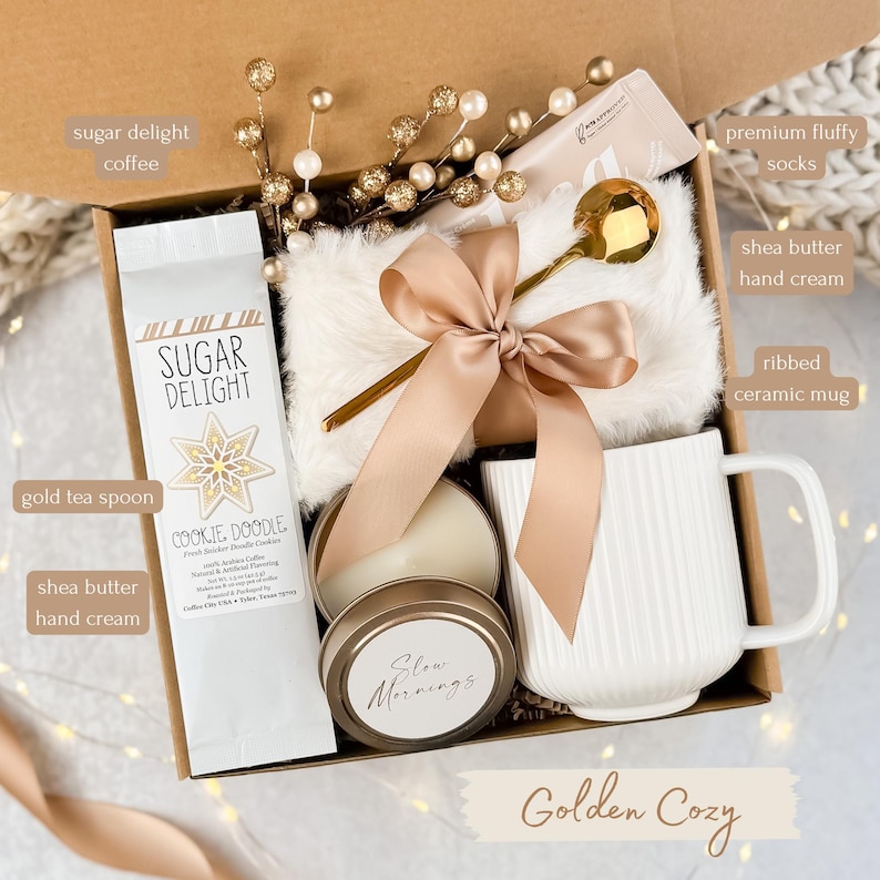 May include: A gift box with a cozy theme. It contains a white ribbed ceramic mug, a gold tea spoon, a candle, a bag of coffee, a pair of fluffy socks, and shea butter hand cream. The box is tied with a satin bow.