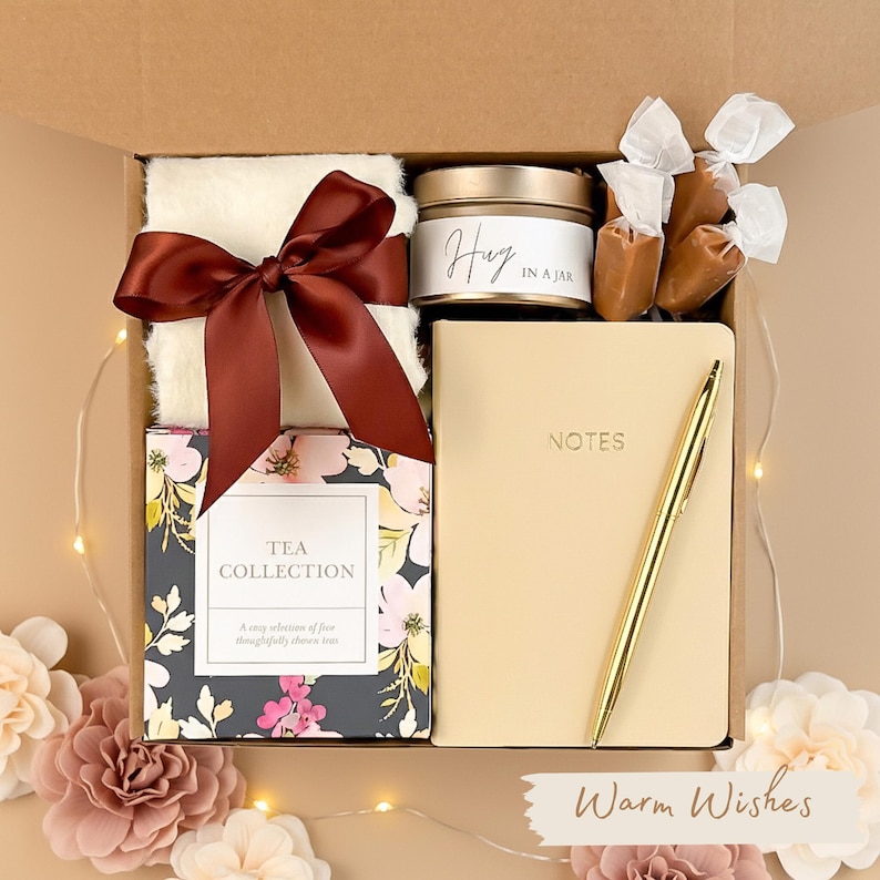 May include: A gift box filled with a cream-colored throw blanket tied with a burgundy bow, a candle labeled "Hug in a Jar", a notebook, a gold pen, tea, and wrapped caramels. The box is decorated with flowers and the words "Warm Wishes".