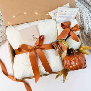 May include: A gift box containing a white diamond-patterned knit blanket, a Moonstruck chocolate bar wrapped in a copper ribbon, two Tea With Tae tea packets, a small candle, a bundle of cinnamon sticks, and a ceramic orange pumpkin figurine.