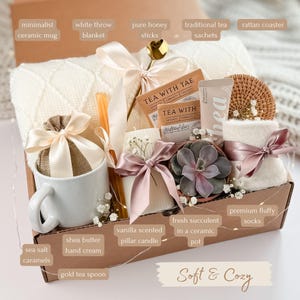 May include: A gift box filled with a white throw blanket, a ceramic mug, tea sachets, a vanilla-scented candle, and a succulent in a ceramic pot. The box also includes honey sticks, a gold tea spoon, shea butter hand cream, and fluffy socks. The words "Soft & Cozy" are written on the box.