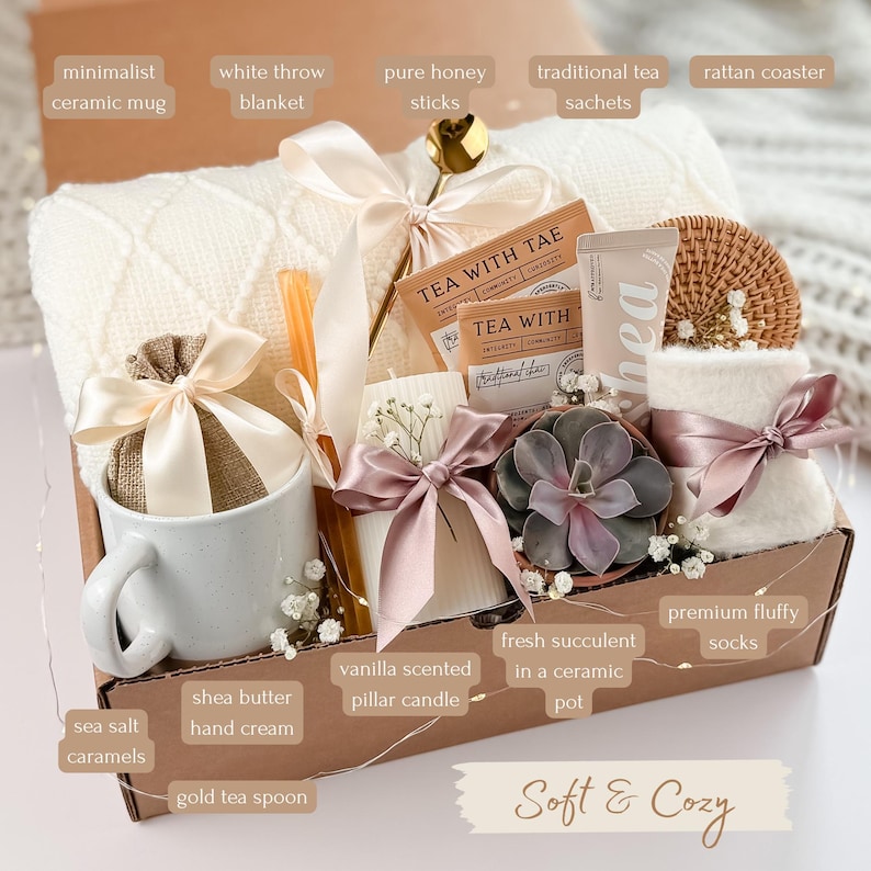 May include: A gift box filled with a white throw blanket, a ceramic mug, tea sachets, a vanilla-scented candle, and a succulent in a ceramic pot. The box also includes honey sticks, a gold tea spoon, shea butter hand cream, and fluffy socks. The words "Soft & Cozy" are written on the box.