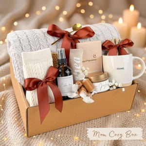 Puede incluir: Una caja de regalo llena de art&iacute;culos acogedores, que incluyen una manta blanca de punto de cable, calcetines, una taza con "mam&aacute;" impresa, una vela y un frasco rociador. La caja est&aacute; atada con cintas color burdeos e incluye el texto "Mom Cozy Box".