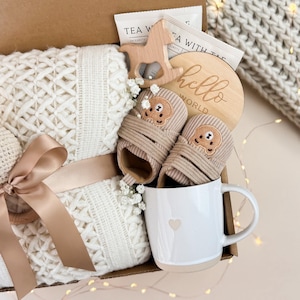 May include: A gift box filled with baby items. Includes a cream-colored knitted blanket with a satin ribbon, a pair of tan baby booties, a wooden teether, a mug with a heart, and a wooden disc with "hello world" text.