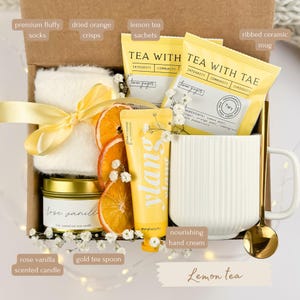 May include: A gift box with a variety of items, including a white ribbed ceramic mug, lemon tea sachets, a rose vanilla scented candle, dried orange crisps, and a tube of nourishing hand cream. The box also contains a gold tea spoon and premium fluffy socks.