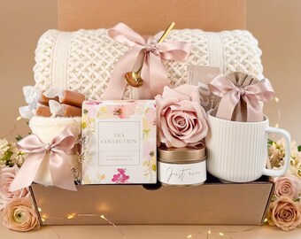 May include: A gift box filled with a white knitted blanket, a tea collection box, a white mug, a candle, and a pink rose. The box is tied with a pink ribbon and includes a gold spoon and wrapped candies. The overall theme is a cozy gift set.