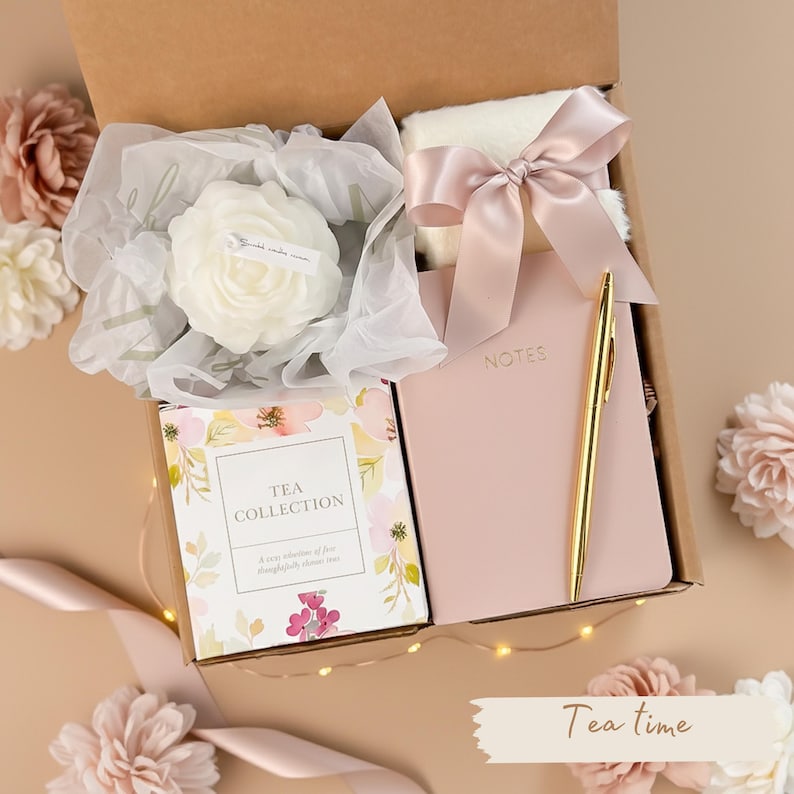 May include: A gift box filled with a white rose-shaped candle, a pink notebook, a gold pen, a tea collection box, and a fluffy white blanket tied with a pink ribbon. The box is decorated with flowers and the words "Tea time".