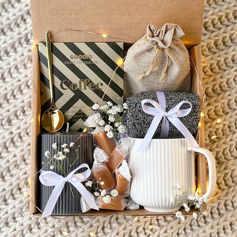 May include: A gift box filled with coffee-themed items. Includes a chocolate bar, a gold spoon, a gray candle, wrapped caramels, a white mug, a small burlap bag, and a gray knit item, all tied with white ribbons and fairy lights.