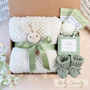 May include: A baby gift box with a cream-colored knitted blanket, a crochet bunny, and a sage green ribbon. The box also contains baby booties, tea bags, and a box of salted caramels. The words "Baby Serenity" are written on the bottom.
