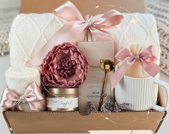 May include: A gift box filled with self-care items, including a white blanket, a pink flower, a candle that says "I am Joyful", a mug, a bag of hot chocolate, and a gold spoon. The box is labelled "Peony Charm".