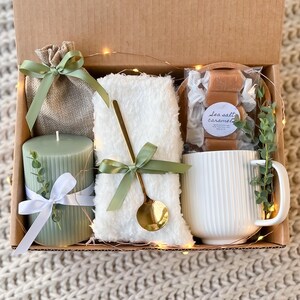 May include: A gift box filled with a green candle, a white mug, a white fluffy blanket, a small burlap bag, and a bag of sea salt caramels. The box is decorated with green and white ribbon and a sprig of greenery.