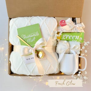 Puede incluir: Una caja de regalo llena de una manta blanca de tejido de gofre, una taza, una barra de chocolate y una barra de t&eacute; verde. La caja est&aacute; atada con una cinta color crema e incluye una tarjeta que dice "Chamomile Mint" y una cuchara dorada. El texto "Fresh Glow" est&aacute; en la parte inferior.