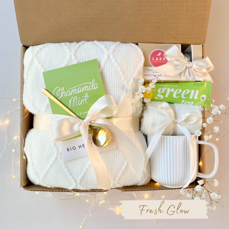 Puede incluir: Una caja de regalo llena de una manta blanca de tejido de gofre, una taza, una barra de chocolate y una barra de t&eacute; verde. La caja est&aacute; atada con una cinta color crema e incluye una tarjeta que dice "Chamomile Mint" y una cuchara dorada. El texto "Fresh Glow" est&aacute; en la parte inferior.