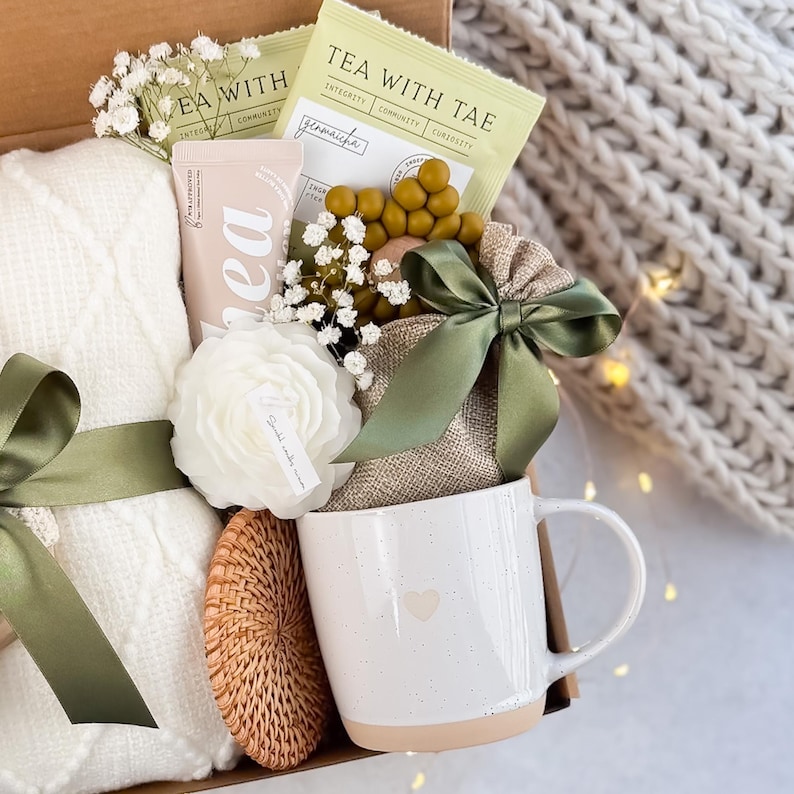 May include: A gift box filled with a white cable-knit blanket, a white mug with a heart, tea packets, a candle, and a woven coaster. The box is decorated with a green ribbon and sprigs of white flowers.