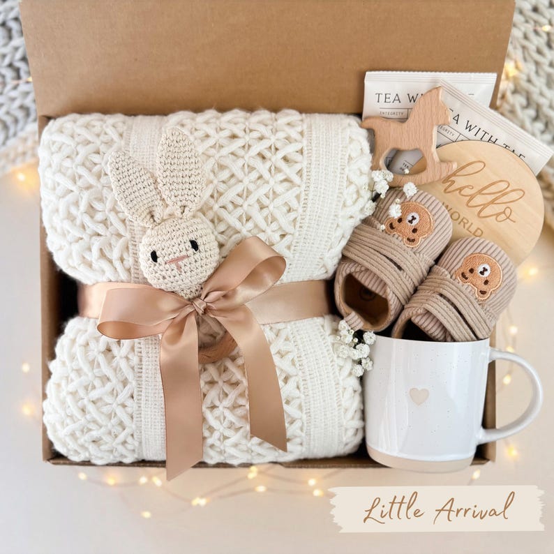 May include: A baby gift set in a brown box, featuring a cream-colored knitted blanket with a crochet bunny, a pair of baby shoes, a wooden teether, a mug, and a wooden disc with the words "hello world". The words "Little Arrival" are at the bottom.