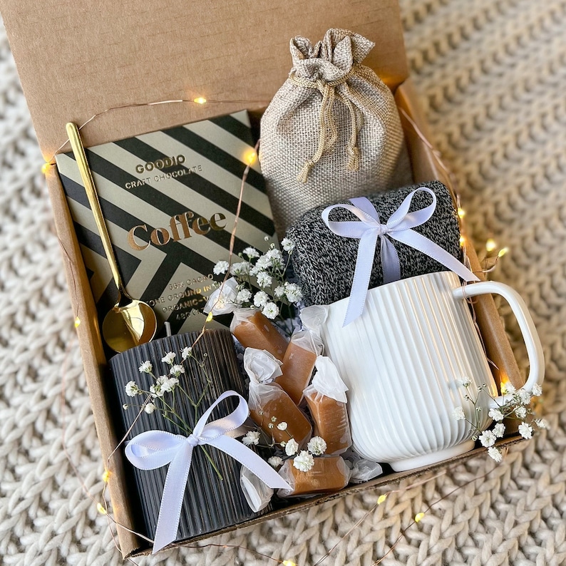 May include: A gift box filled with a white mug, a black candle, a chocolate bar, a gold spoon, a small burlap bag, a pair of gray socks, and wrapped caramels. The box is decorated with white flowers and a string of fairy lights.