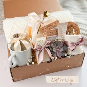 Puede incluir: Una caja de regalo llena de art&iacute;culos acogedores. Incluye una taza blanca, una peque&ntilde;a bolsa atada con una cinta, sobres de t&eacute;, una vela, una suculenta, una toalla peque&ntilde;a y un posavasos tejido. Tambi&eacute;n se incluye una manta color crema y una cuchara dorada. La caja est&aacute; atada con cintas.