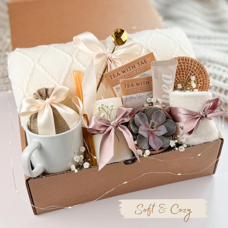 Puede incluir: Una caja de regalo llena de art&iacute;culos acogedores. Incluye una taza blanca, una peque&ntilde;a bolsa atada con una cinta, sobres de t&eacute;, una vela, una suculenta, una toalla peque&ntilde;a y un posavasos tejido. Tambi&eacute;n se incluye una manta color crema y una cuchara dorada. La caja est&aacute; atada con cintas.