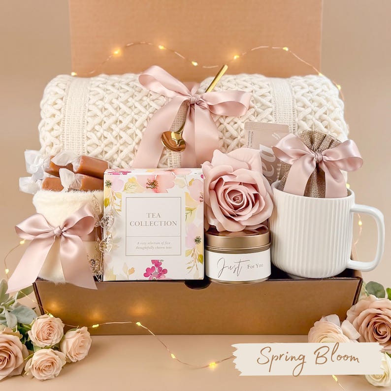 Puede incluir: Una caja de regalo llena de una manta de punto color crema, una taza blanca, una caja de colecci&oacute;n de t&eacute;, una vela, una rosa y otros art&iacute;culos. La caja est&aacute; atada con cintas rosas e incluye el texto "Spring Bloom".