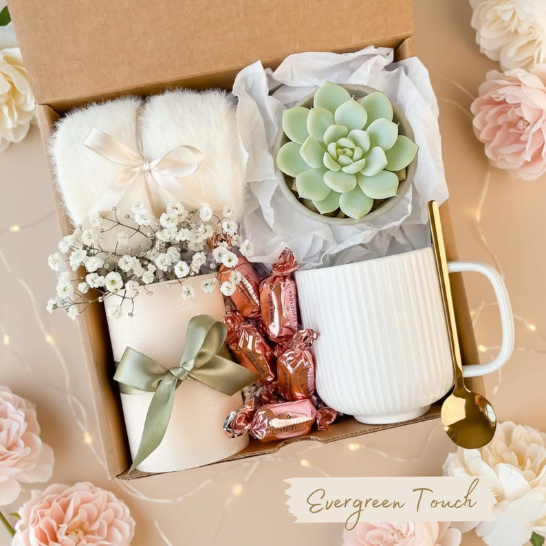 May include: A gift box filled with a succulent in a gray pot, a white mug, a gold spoon, a pair of fluffy slippers, a candle with a green bow, and wrapped candies. The box is lined with white tissue paper and has the text "Evergreen Touch".