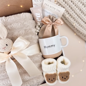 May include: A gift box filled with a white mug with "Mommy" printed on it, a plush blanket, baby booties, tea packets, and a small bag tied with a bow. The booties have a brown bear face design.