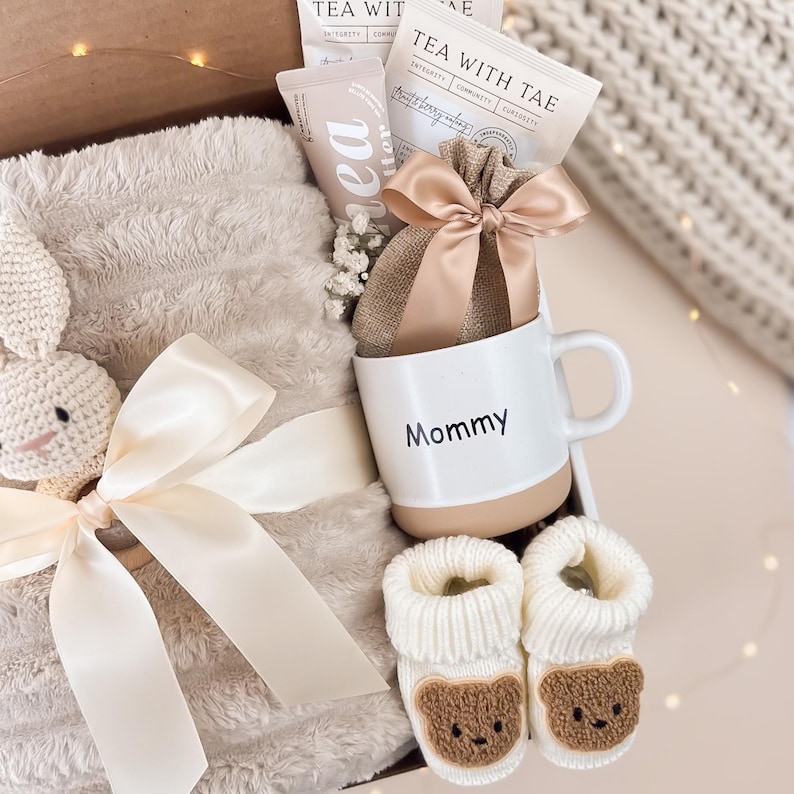 May include: A gift box filled with a white mug with "Mommy" printed on it, a plush blanket, baby booties, tea packets, and a small bag tied with a bow. The booties have a brown bear face design.