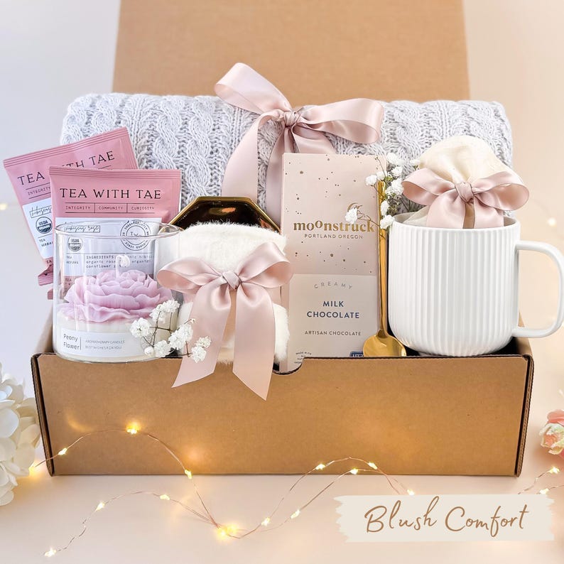 Puede incluir: Una caja de regalo llena de una manta de punto trenzado gris, sobres de t&eacute;, una vela, una taza y chocolate. Los art&iacute;culos est&aacute;n atados con cintas rosas. La caja tiene el texto "Blush Comfort".