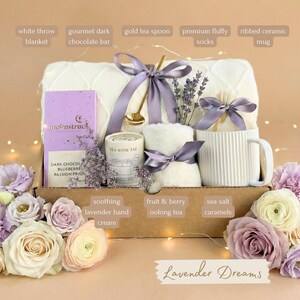 May include: A gift box with a white throw blanket, gourmet dark chocolate bar, gold tea spoon, premium fluffy socks, and a ribbed ceramic mug. The box also contains lavender hand cream, fruit & berry oolong tea, and sea salt caramels. The text "Lavender Dreams" is visible.