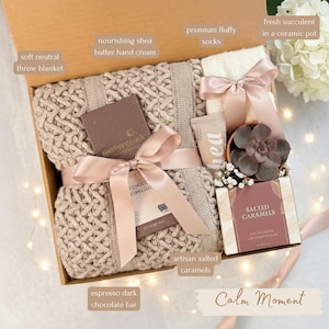 May include: A gift box with a soft neutral throw blanket, premium fluffy socks, and artisan salted caramels. Includes a dark chocolate bar, shea butter hand cream, and a succulent in a ceramic pot. The box is tied with a pink ribbon.