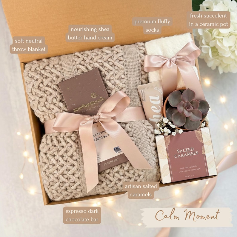 May include: A gift box with a soft neutral throw blanket, premium fluffy socks, and artisan salted caramels. Includes a dark chocolate bar, shea butter hand cream, and a succulent in a ceramic pot. The box is tied with a pink ribbon.