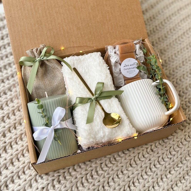 May include: A gift box filled with self-care items, including a white towel, a green candle, a mug, and a bag of sea salt caramel sweets. The box is decorated with green ribbon and a sprig of greenery.