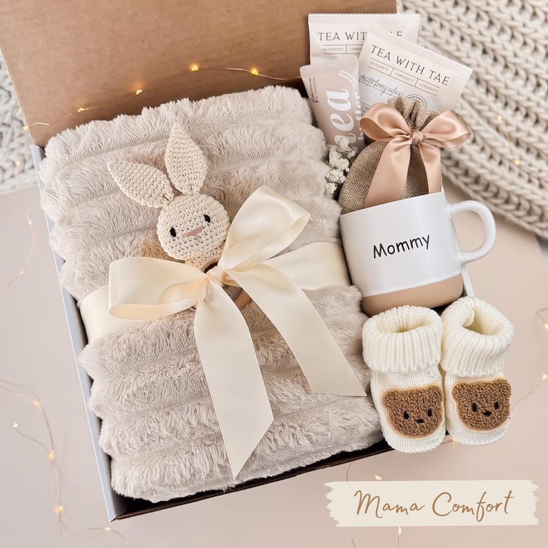 May include: A gift box filled with a plush blanket, a crocheted bunny, a mug with "Mommy" printed on it, tea bags, and baby booties. The blanket is beige and tied with a cream ribbon. The booties have bear faces.