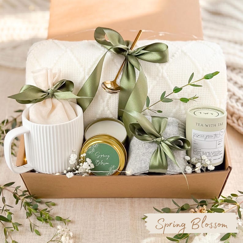 Puede incluir: Una cesta de regalo llena de una taza blanca, una vela peque&ntilde;a, un bote de t&eacute; y una manta doblada. Los art&iacute;culos est&aacute;n atados con cintas verde oliva y colocados en una caja marr&oacute;n. Las palabras "Spring Blossom" est&aacute;n escritas en una pancarta.