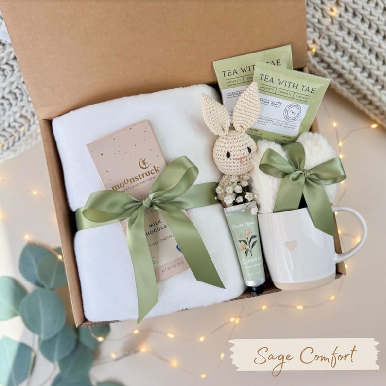 May include: A gift box filled with a white towel, chocolate bar, tea packets, a crocheted bunny, hand cream, and a mug. The items are tied with sage green ribbons. The box is labeled "Sage Comfort".