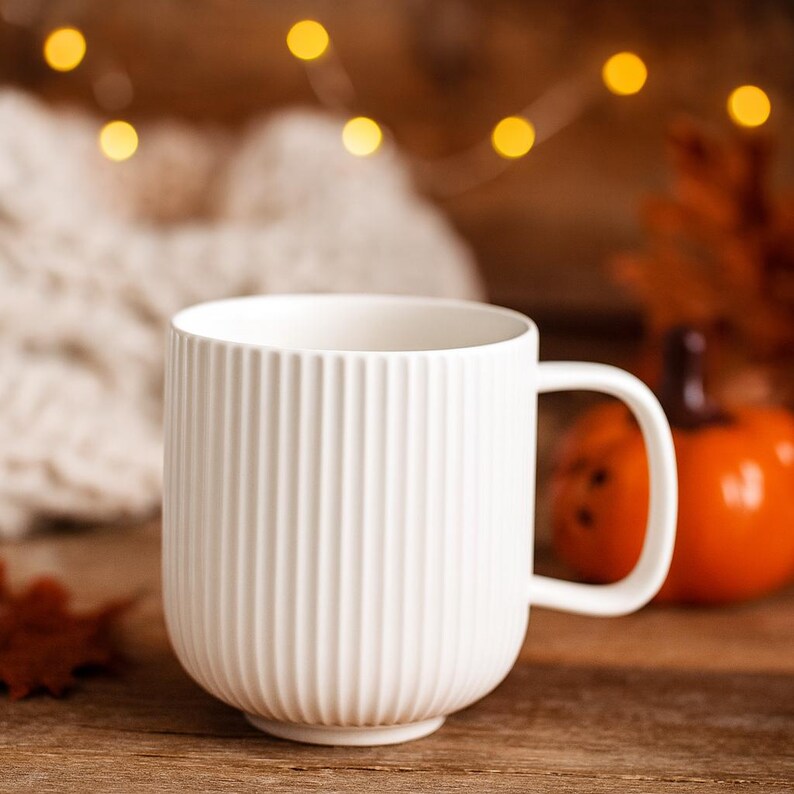 May include: A white ceramic mug with vertical ribbed detailing. The mug has a curved handle and sits on a wooden surface. The background includes a blurred knitted item, a small orange pumpkin, and string lights.