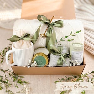 May include: A gift box with a white mug, a small candle, a tea container, and a gray item, all tied with green ribbons. A white blanket and a gold spoon are also included. The box is labeled "Spring Blossom".