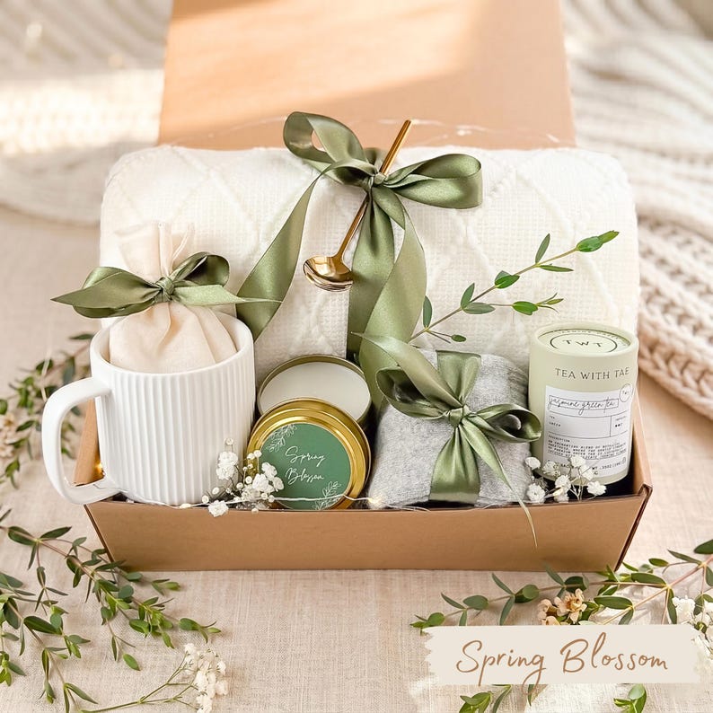 May include: A gift box with a white mug, a small candle, a tea container, and a gray item, all tied with green ribbons. A white blanket and a gold spoon are also included. The box is labeled "Spring Blossom".