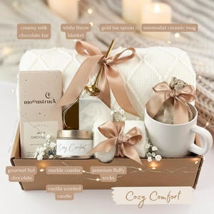 May include: A gift basket filled with a white throw blanket, a minimalist ceramic mug, and a gold tea spoon. The basket also contains a creamy milk chocolate bar, gourmet hot chocolate, and a vanilla scented candle. The text "Cozy Comfort" is visible.