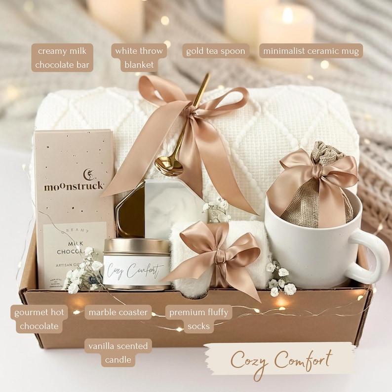 May include: A gift basket filled with a white throw blanket, a minimalist ceramic mug, and a gold tea spoon. The basket also contains a creamy milk chocolate bar, gourmet hot chocolate, and a vanilla scented candle. The text "Cozy Comfort" is visible.