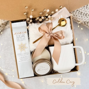 May include: A gift box filled with a white mug, a gold spoon, a candle, a bag of coffee, and a fluffy white blanket. The box is tied with a gold ribbon. The coffee bag reads "Sugar Delight Cookie Doodle".