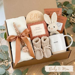 May include: A gift box filled with baby and mom essentials. Includes a muslin blanket, wooden teether, knitted booties, a mug with "mom" printed on it, a crochet bunny, salted caramels, and tea. The box has the text "hello world".