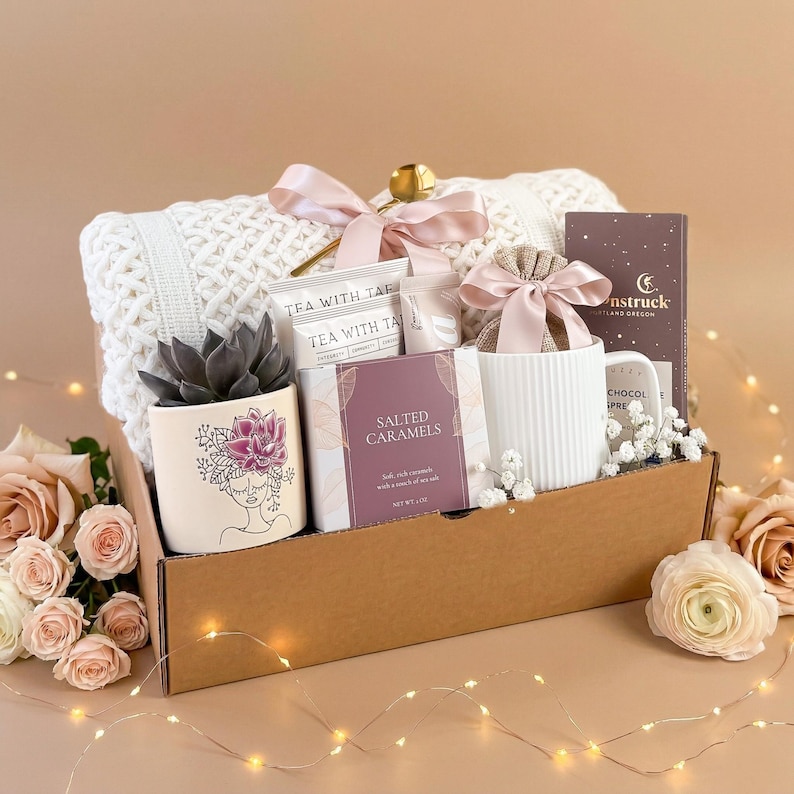 Pu&ograve; includere: Un cesto regalo pieno di una coperta bianca lavorata a maglia, una pianta succulenta in un vaso decorativo, bustine di t&egrave;, caramelle al sale, una tazza e una tavoletta di cioccolato. Il cesto &egrave; legato con un nastro rosa e si trova in una scatola di cartone marrone.