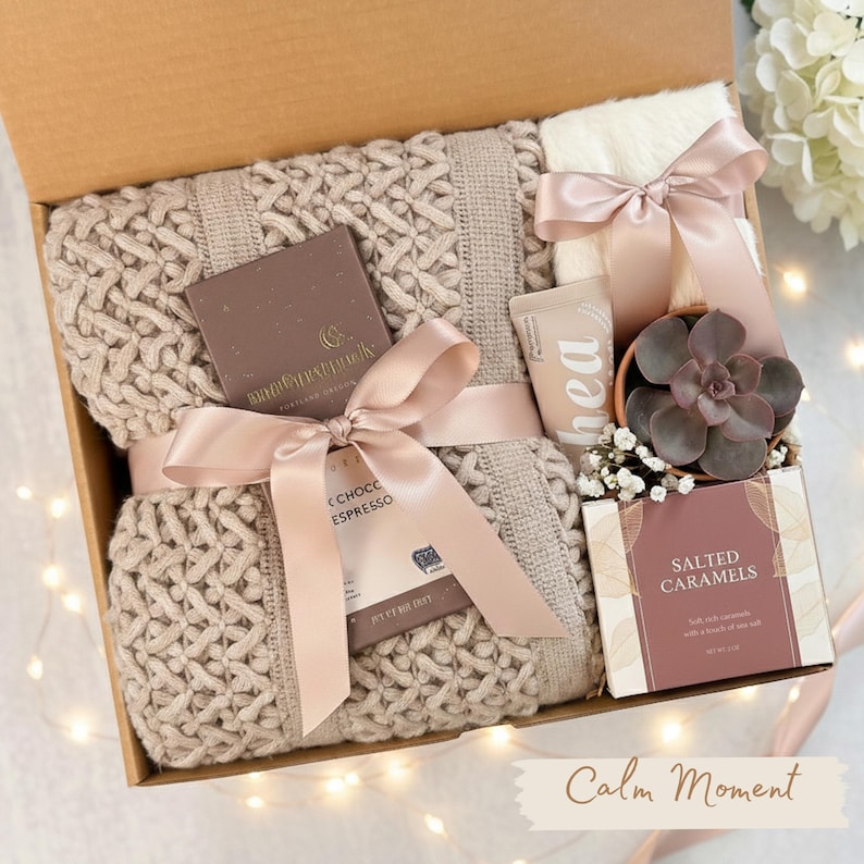 Puede incluir: Una caja de regalo llena de una manta de punto beige, chocolate, una peque&ntilde;a suculenta en una maceta, un tubo de loci&oacute;n y un art&iacute;culo blanco esponjoso, todo atado con una cinta rosa. La caja tambi&eacute;n contiene una caja de caramelos de sal. El texto "Calm Moment" est&aacute; en la parte inferior.